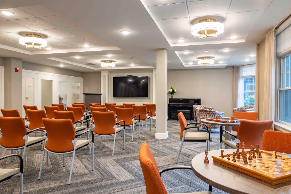 A spacious activity room with rows of orange chairs facing a large flat-screen TV mounted on the wall. The room has modern ceiling lights, a carpeted floor with a geometric pattern, and large windows with beige curtains letting in natural light. In the foreground, there is a round table with a chessboard set up, and in the background, there is a black piano and additional seating with patterned armchairs.