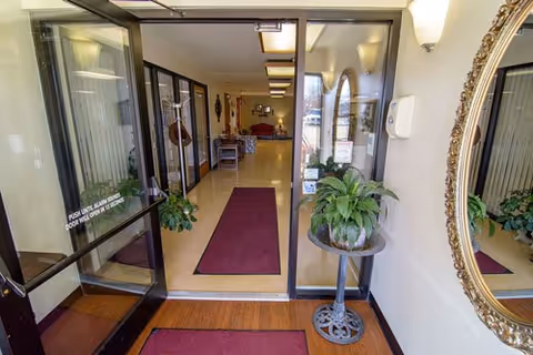 Glass entrance doors opening into a lobby hallway with maroon runners, potted plants, and seating visible down the corridor.
