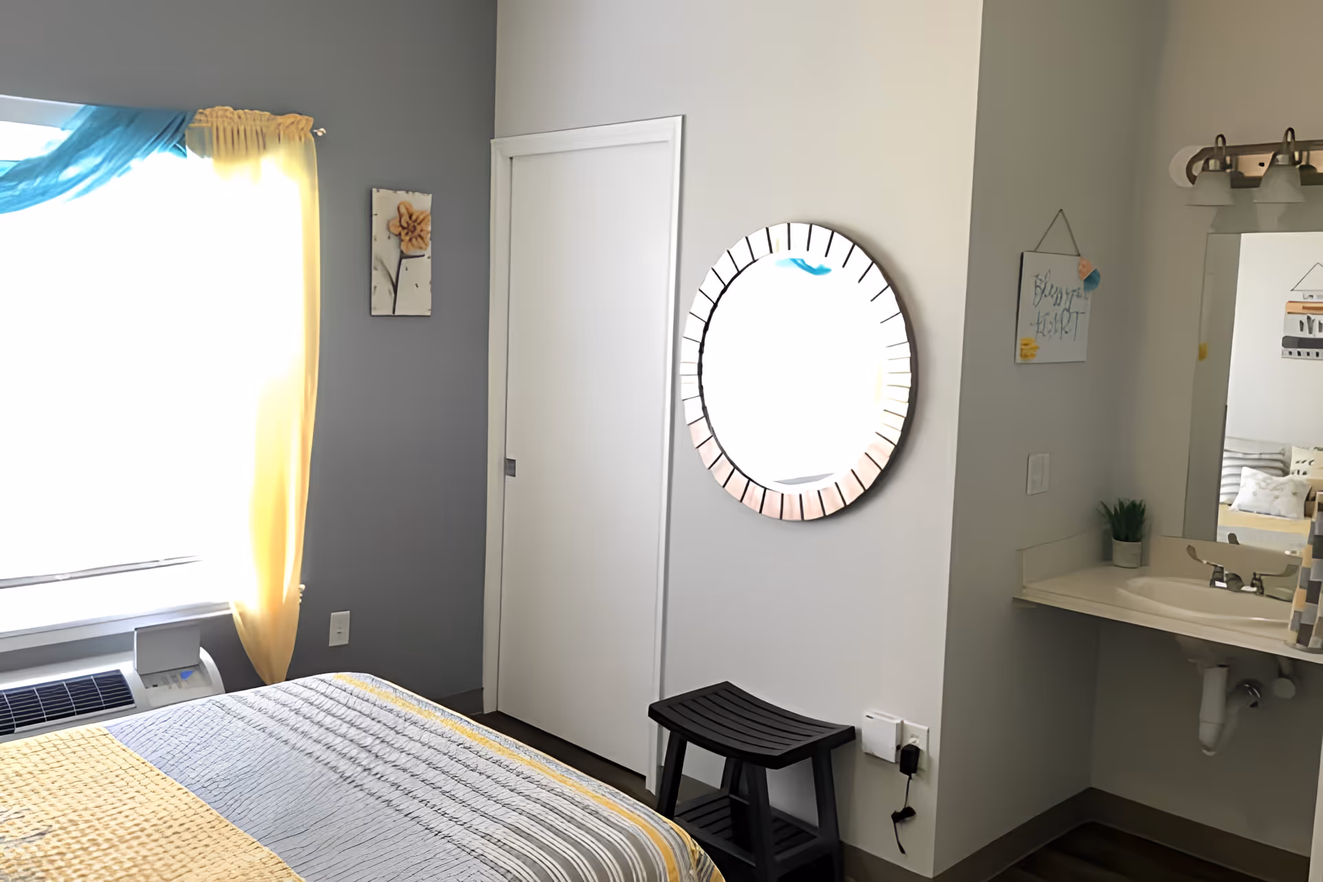 A bedroom with a bed covered in a gray and yellow quilt, a window with yellow and blue curtains, a white sliding closet door, a round mirror on the wall, a small black step stool, and a sink with a mirror and light fixture above it.