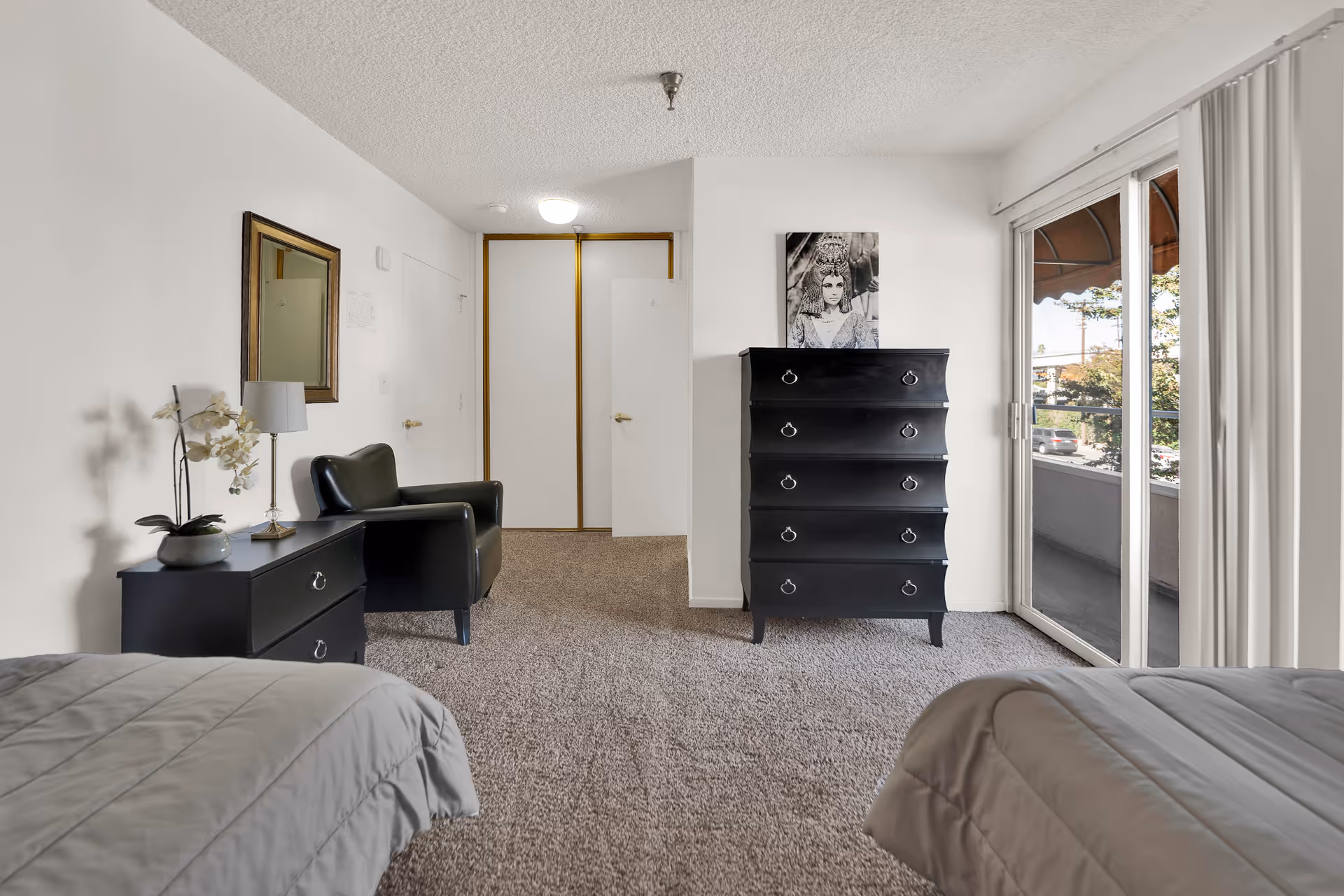 A bedroom with two beds partially visible on either side, a black armchair, a black nightstand with a lamp and a potted plant, a black chest of drawers with a black and white portrait above it, beige carpet, white walls, and a sliding glass door leading to a balcony with a view of trees and parked cars outside.