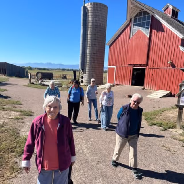 A group of six elderly people walking outdoors on a farm path near a large red barn and a tall silo under a clear blue sky.
