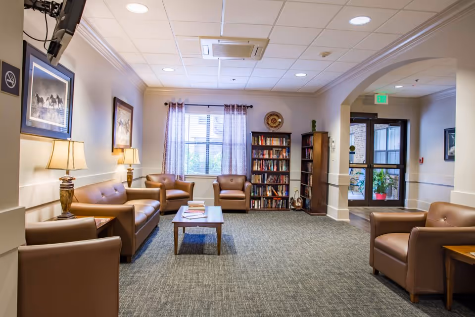 A cozy living room area in a senior living facility with several brown leather chairs and sofas arranged around a wooden coffee table. There are two table lamps on side tables, framed pictures on the walls, a bookshelf filled with books, and a window with sheer curtains letting in natural light. The room has a carpeted floor and a ceiling with recessed lighting and an air conditioning unit. A glass door leads to another area with visible plants outside.