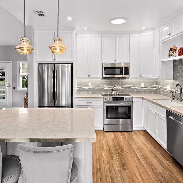 Modern kitchen with white cabinetry, stainless steel refrigerator, oven, microwave, and dishwasher. The kitchen features a light-colored countertop island with two gray cushioned chairs, pendant lights hanging above, and wood flooring. A window and door are visible in the background.