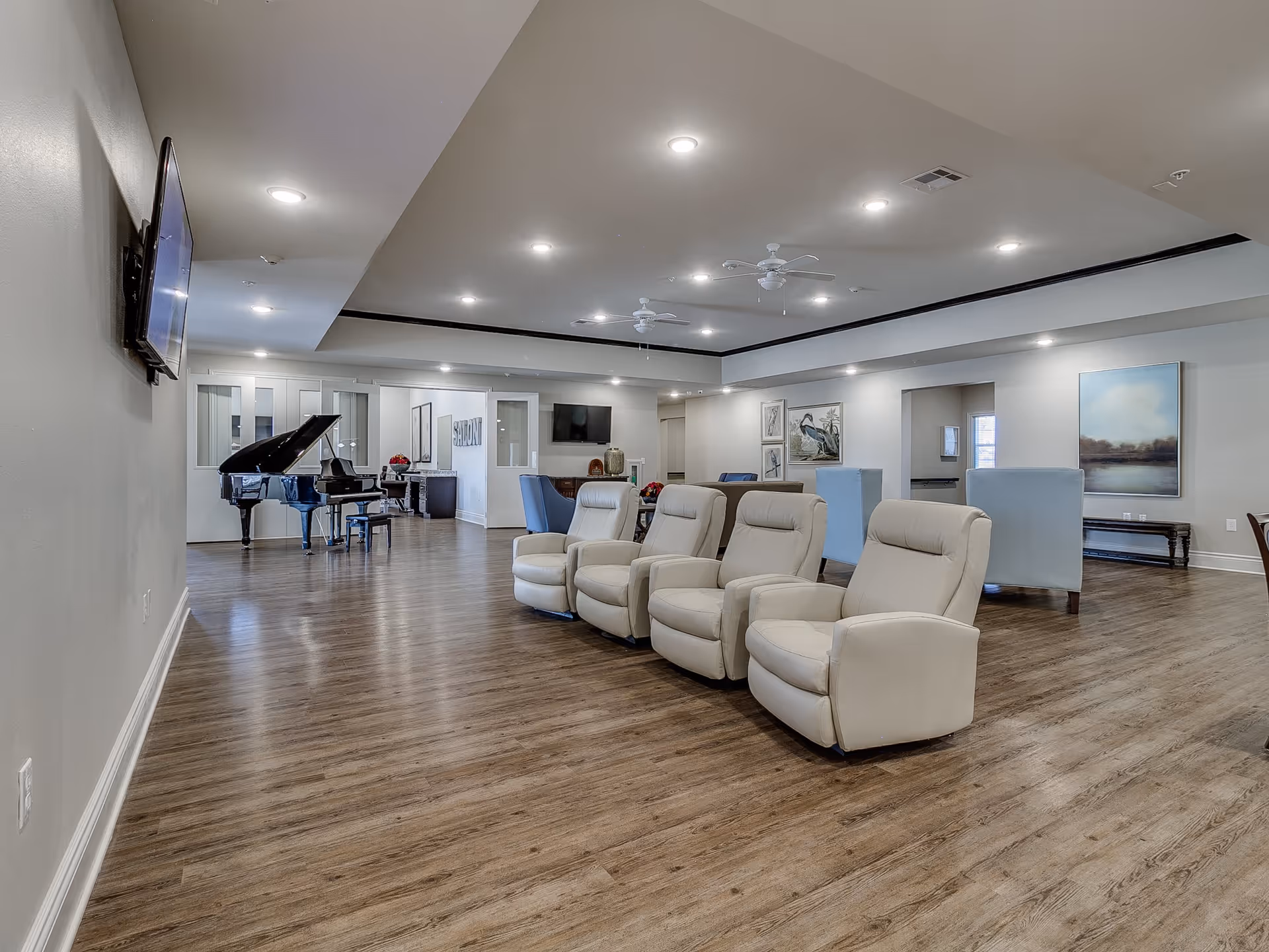 A spacious and well-lit common area with wooden flooring featuring four beige recliner chairs in the foreground, a black grand piano on the left side, a wall-mounted TV, and several blue armchairs and artwork on the walls in the background.