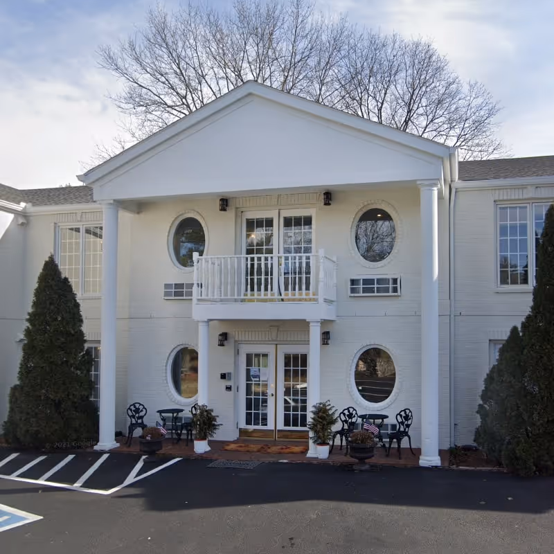 Front exterior view of a two-story white building with a small balcony above the entrance, round windows on either side of the doors, and two large white columns supporting the roof over the entrance. There are outdoor chairs and potted plants near the entrance, and tall evergreen trees on both sides of the building.