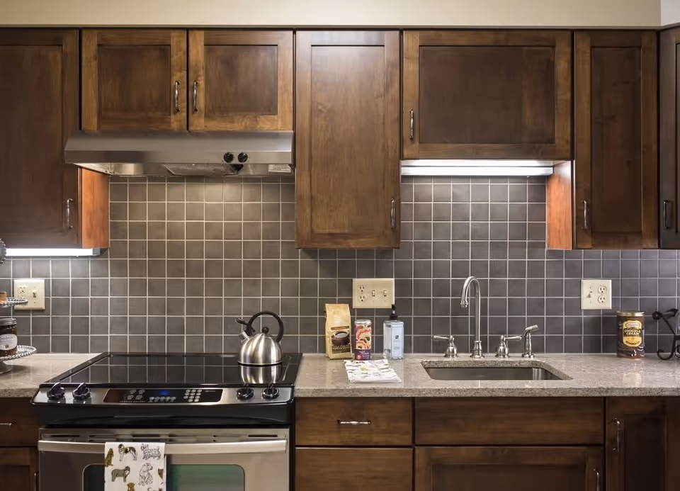 A kitchen with dark wooden cabinets, a stainless steel stove with a kettle on it, a gray tiled backsplash, a stainless steel sink with a faucet, and various kitchen items on the countertop including a coffee bag, hand soap, and a container of cocoa cream.
