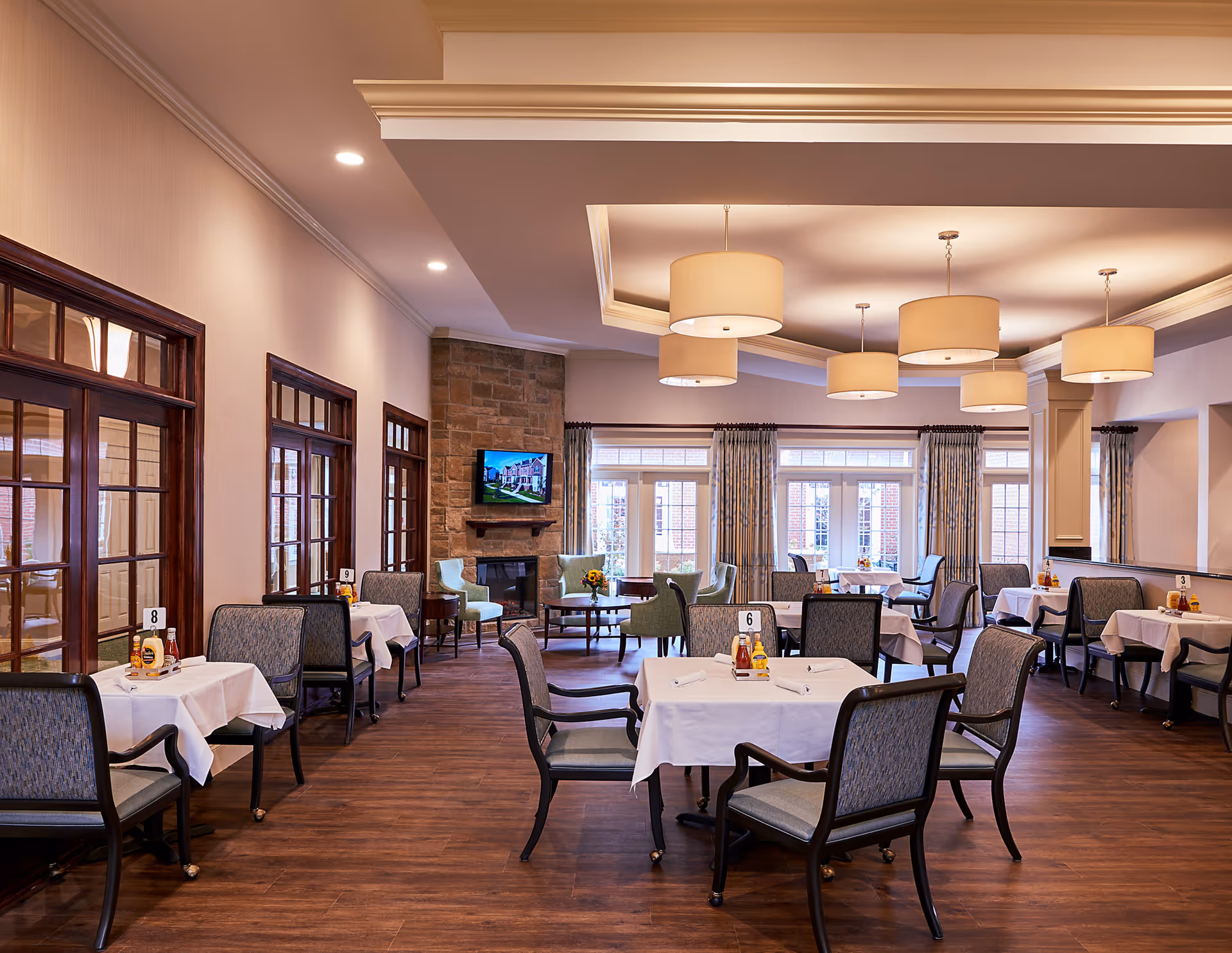 A spacious dining room in a senior living facility with multiple tables covered in white tablecloths, each set with chairs and condiments. The room features large windows with curtains, a stone fireplace with a mounted TV above it, and modern ceiling lights. The floor is wooden, and the walls have wooden framed windows and doors.