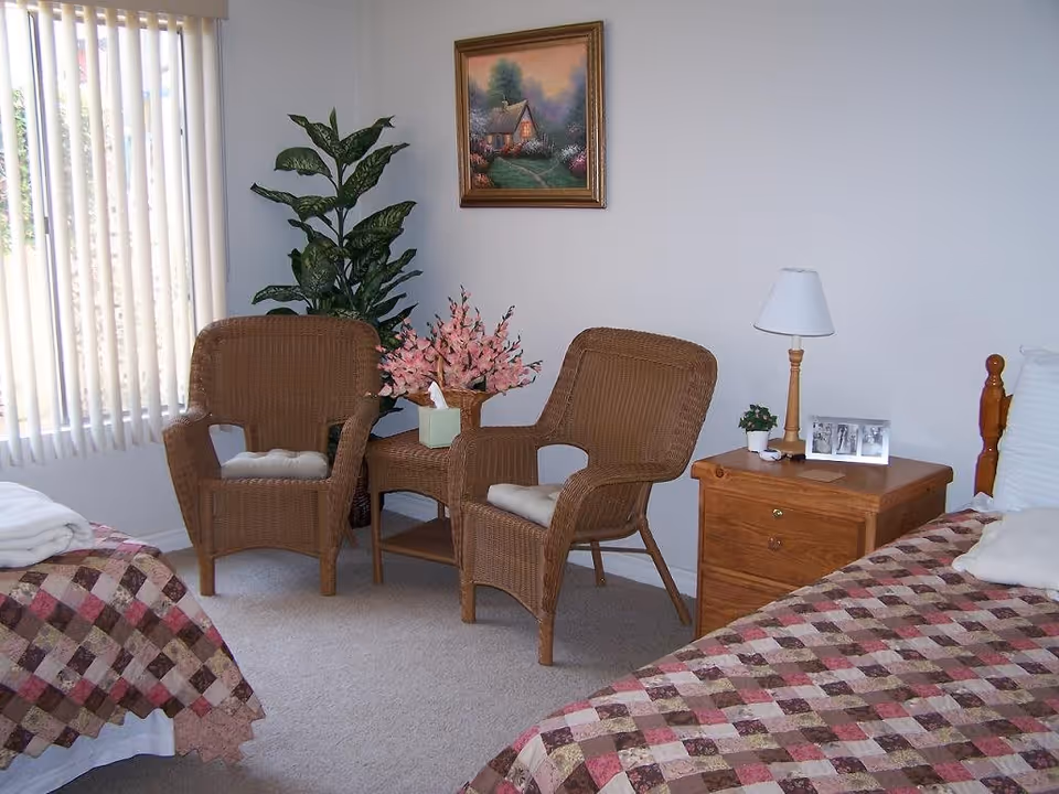A cozy bedroom with two beds covered in patterned quilts, a wooden nightstand with a lamp, a small plant, and a framed photo. Two wicker chairs with cushions and a small wicker table with a tissue box and pink flowers are placed near a window with vertical blinds. A framed painting of a cottage hangs on the wall above the chairs.