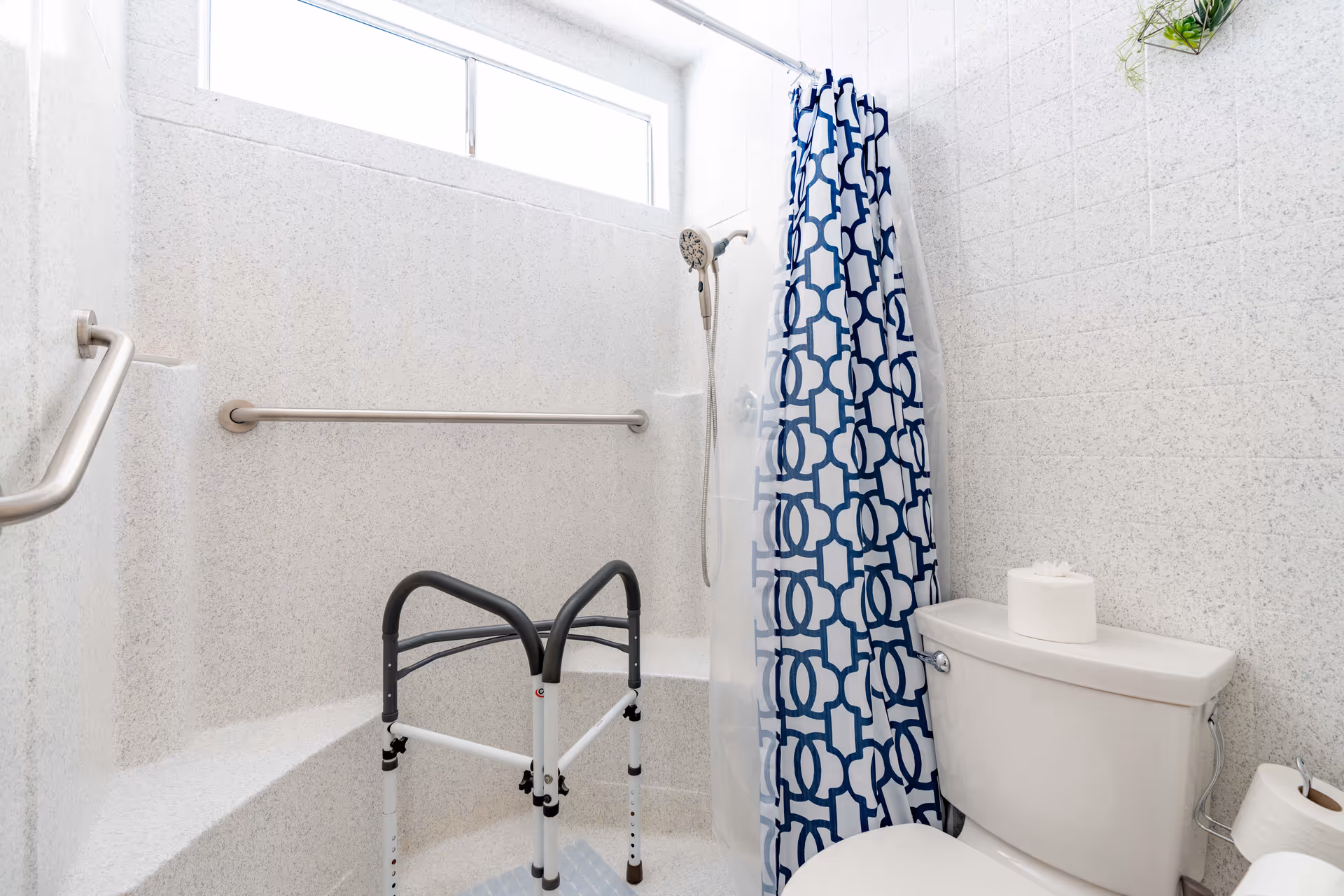 A bathroom with a walk-in shower featuring a built-in bench, grab bars, a handheld showerhead, and a blue and white patterned shower curtain. There is a white toilet with a roll of toilet paper on top and an additional roll on a holder beside it. The walls are light-colored with a speckled texture, and there is a small window above the shower area.