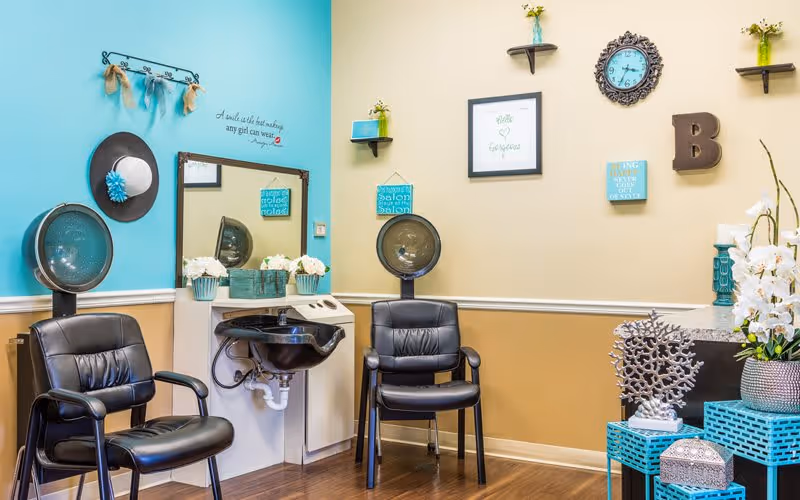 Small salon corner with two black styling chairs, hooded hair dryers, a wash sink, and teal-and-beige wall decor and shelving.