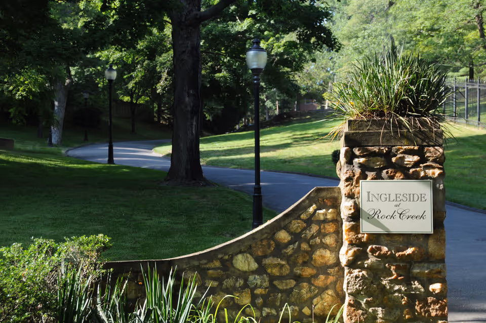 Stone entrance pillar with a sign reading 'Ingleside at Rock Creek' next to a winding paved pathway lined with grass, trees, and lamp posts in a park-like setting.