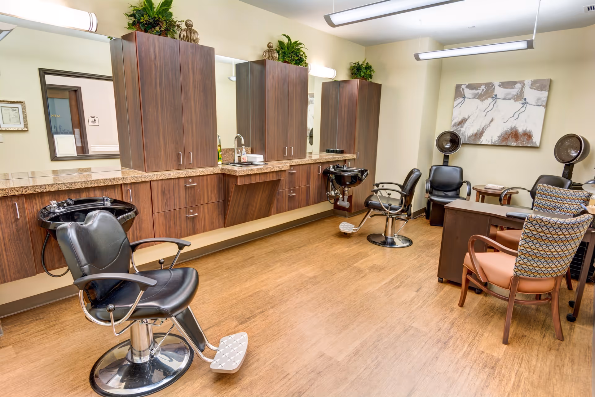 Interior view of a hair salon area in an assisted living facility featuring two black salon chairs with attached sinks, wooden cabinets with granite countertops, a sink, and plants on top of the cabinets. There are also two hair dryers with chairs and a small table with chairs in the corner, along with a painting on the wall.