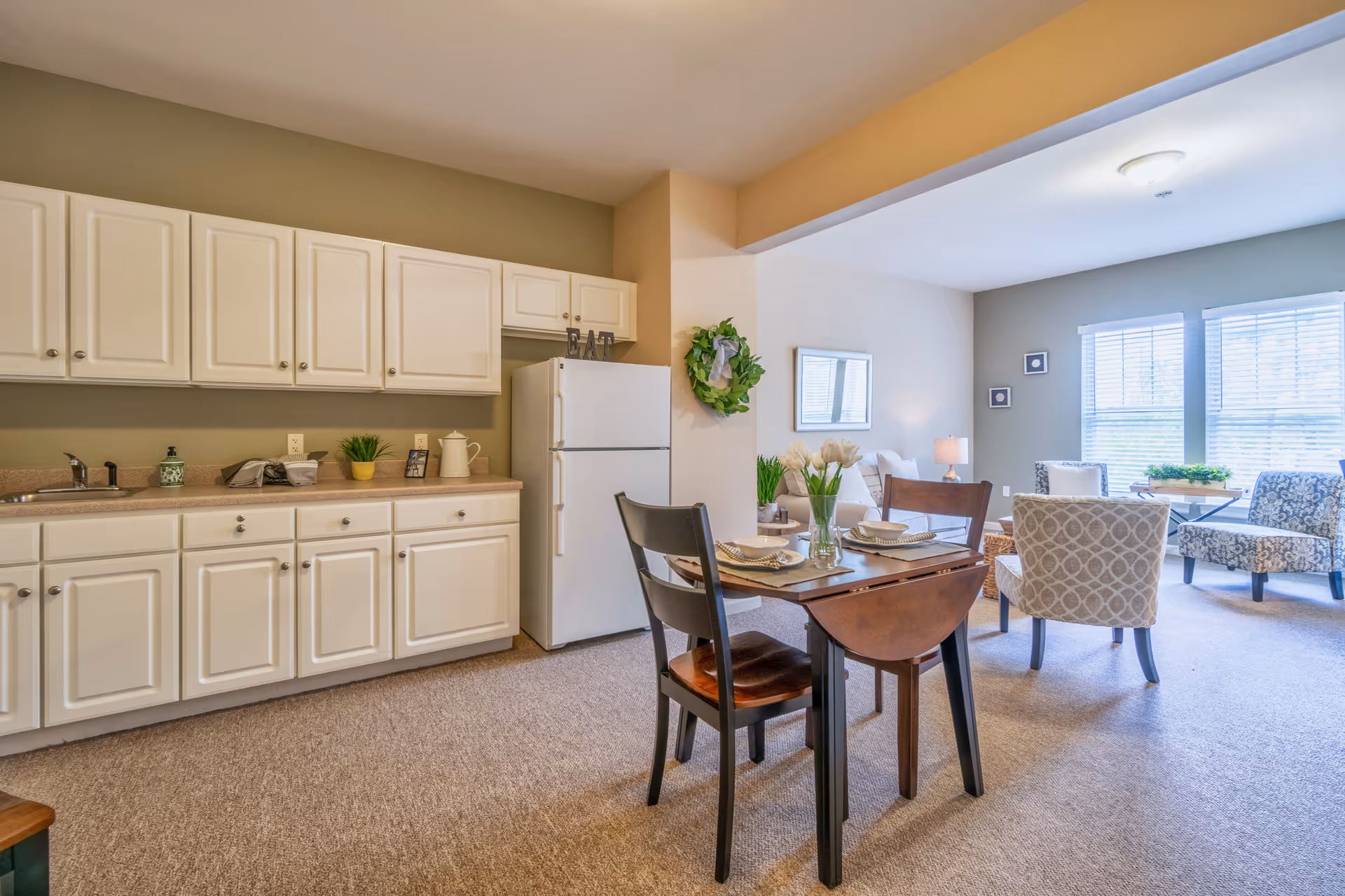 Open-plan senior living apartment interior with a kitchenette of white cabinets and a refrigerator, a small dining table set for two, and a sitting area with chairs and windows.