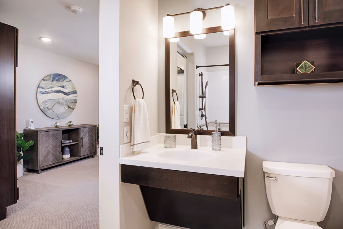 A clean and modern bathroom with a white sink and countertop, a dark wood framed mirror with three lights above it, a towel hanging on a ring, and a white toilet. In the background, there is a glimpse of a room with a wooden cabinet, decorative items, a round abstract wall art, and a potted plant.