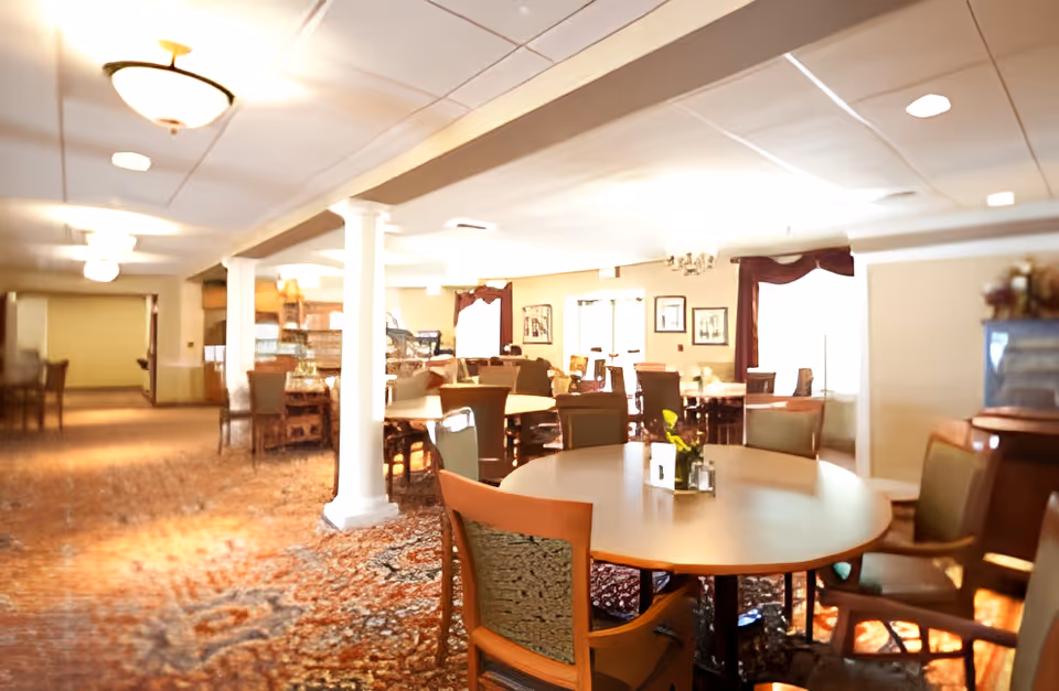 A spacious dining area in a senior living facility with multiple round and rectangular tables surrounded by chairs. The room features carpeted floors with a patterned design, white columns, and soft lighting from ceiling fixtures. The walls are decorated with framed pictures and windows with curtains, creating a warm and inviting atmosphere.