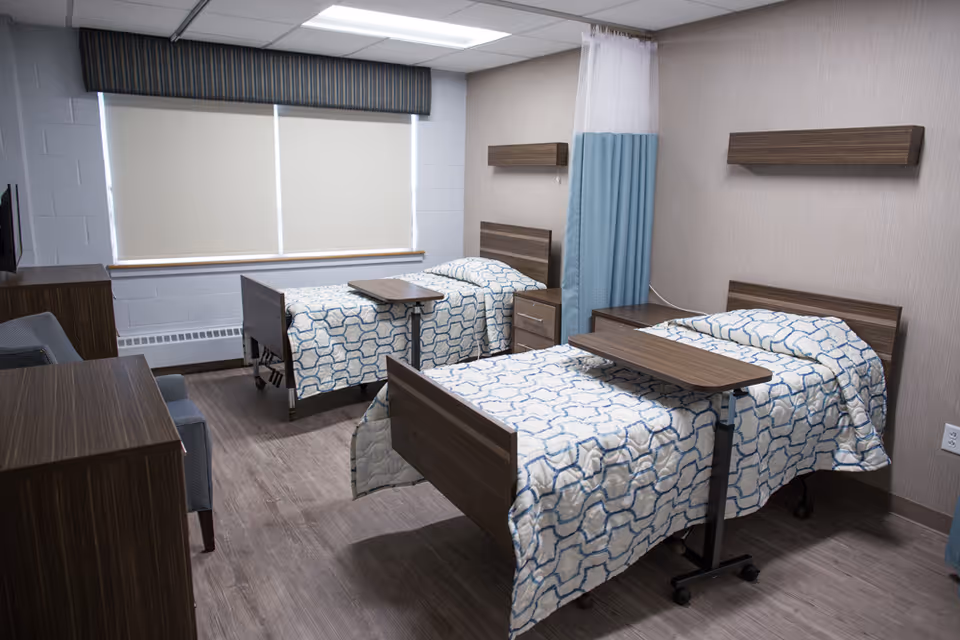 A clean and modern room with two hospital-style beds, each with a patterned blue and white bedspread and an overbed table. The room has wood-look flooring, a window with closed blinds, a blue privacy curtain between the beds, and minimalistic wooden furniture including a dresser and nightstand.