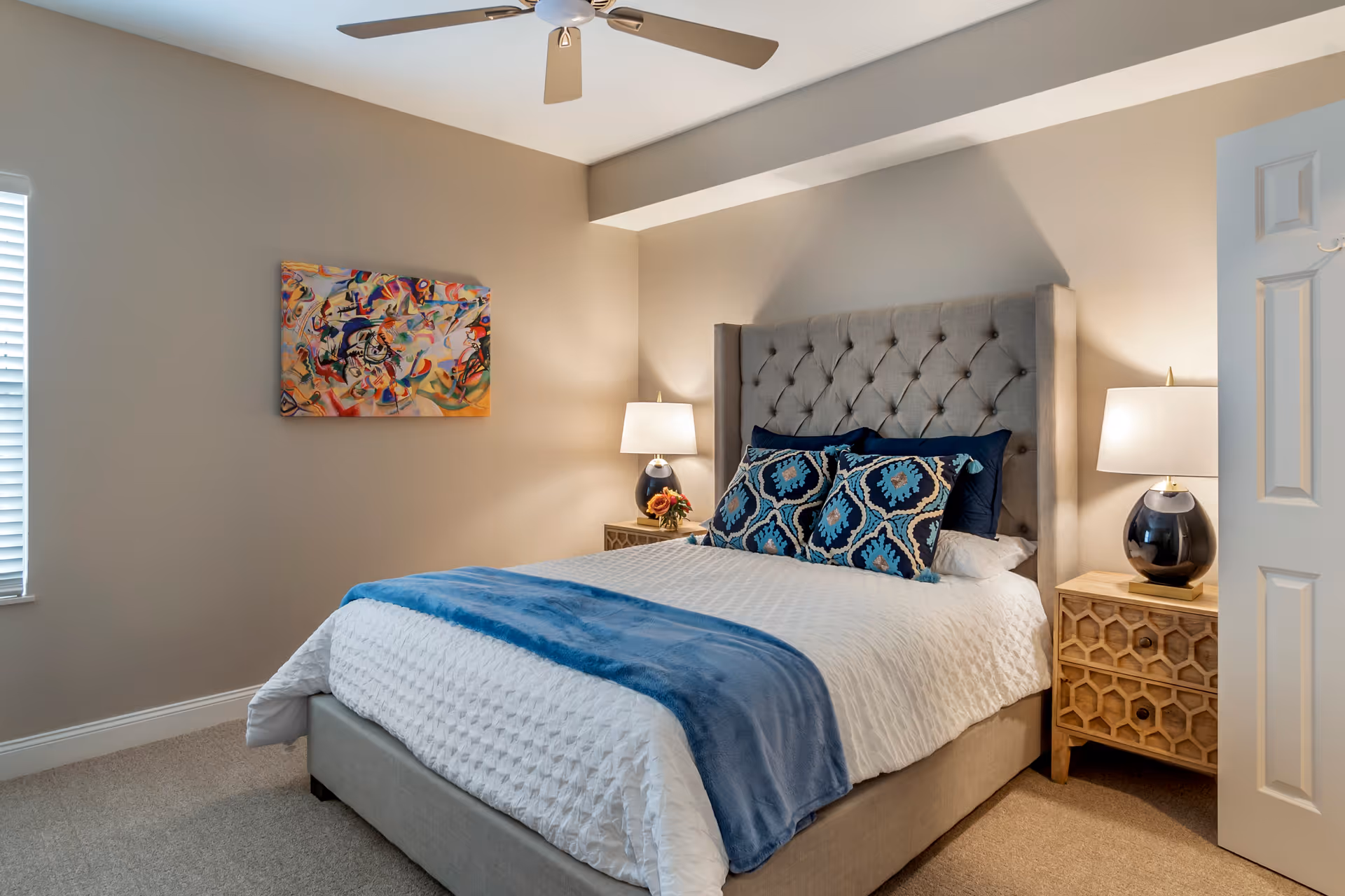 A cozy bedroom with a tufted gray upholstered bed, white bedding, and a blue throw blanket. Two decorative pillows with blue patterns are placed on the bed. There are two wooden nightstands on either side of the bed, each with a lamp featuring a dark base and white shade. A colorful abstract painting hangs on the beige wall to the left, and a ceiling fan is visible above. A window with white blinds is on the left wall, and a partially open white door is on the right.