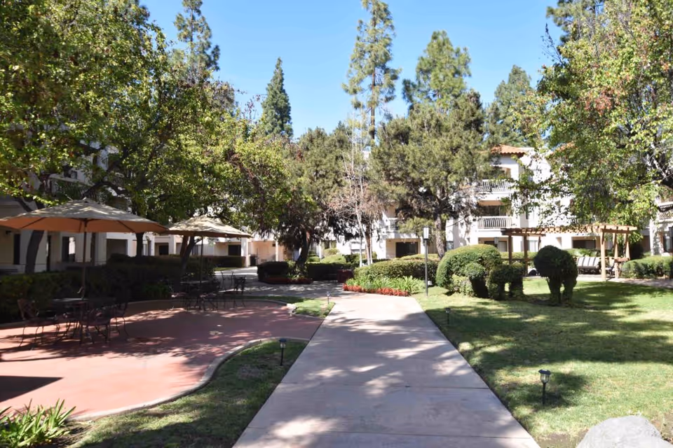 Outdoor courtyard area at Solstice Senior Living at El Cajon featuring a paved walkway, patio with tables and umbrellas, well-maintained green lawn, trees, and shrubbery shaped like animals under a clear blue sky.