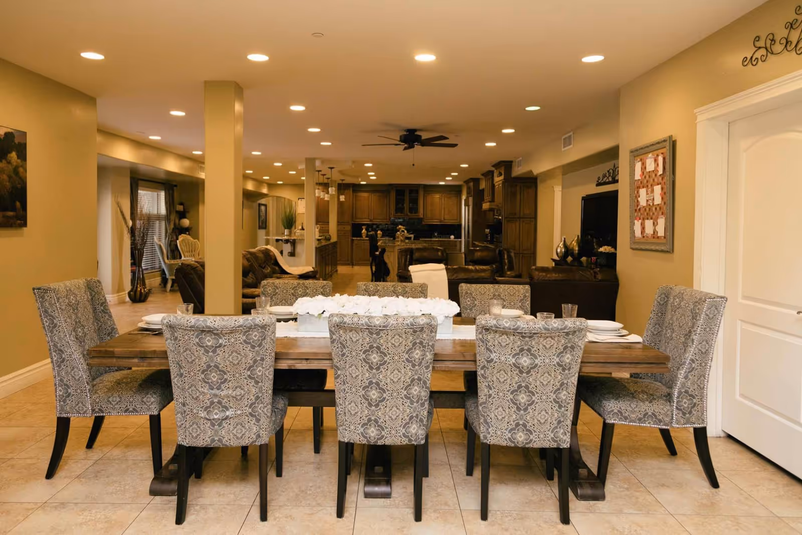 A spacious dining area with a wooden table set for eight, featuring patterned upholstered chairs. The room extends into a living area with leather sofas and a ceiling fan, and a kitchen with wooden cabinets is visible in the background. The walls are painted beige, and the floor is tiled.