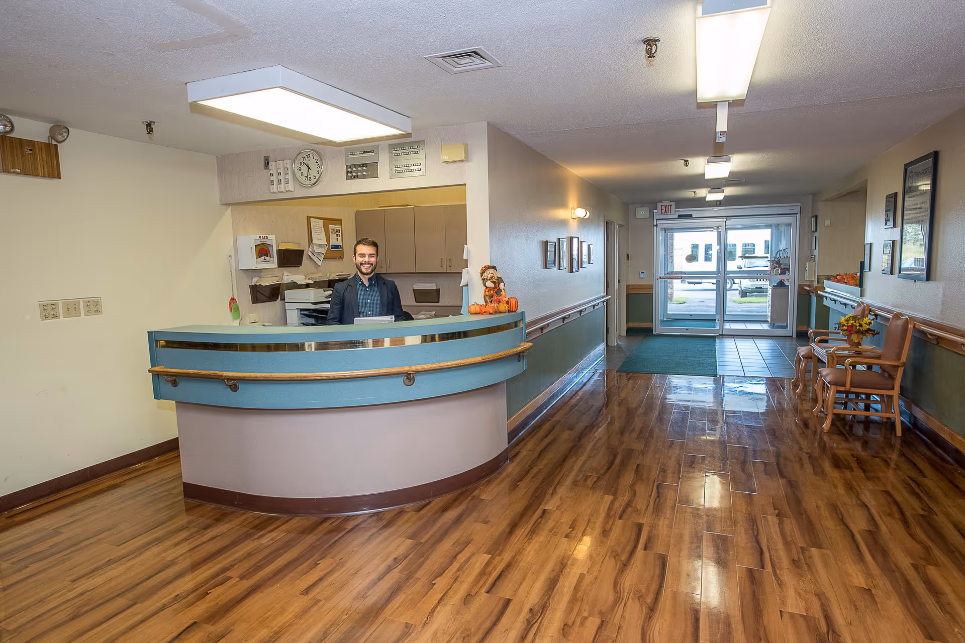 Reception area of Twin Ports Health Services with a smiling receptionist behind a curved desk. The hallway has wooden flooring, chairs with a small table and decorations, and glass doors leading outside.