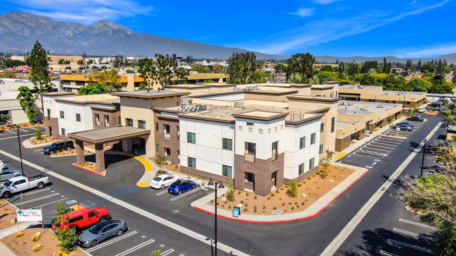 Aerial view of Aqua Ridge of Montclair senior living facility showing a modern two-story building with a covered entrance, surrounded by parking lots and landscaping, with mountains in the background under a clear blue sky.