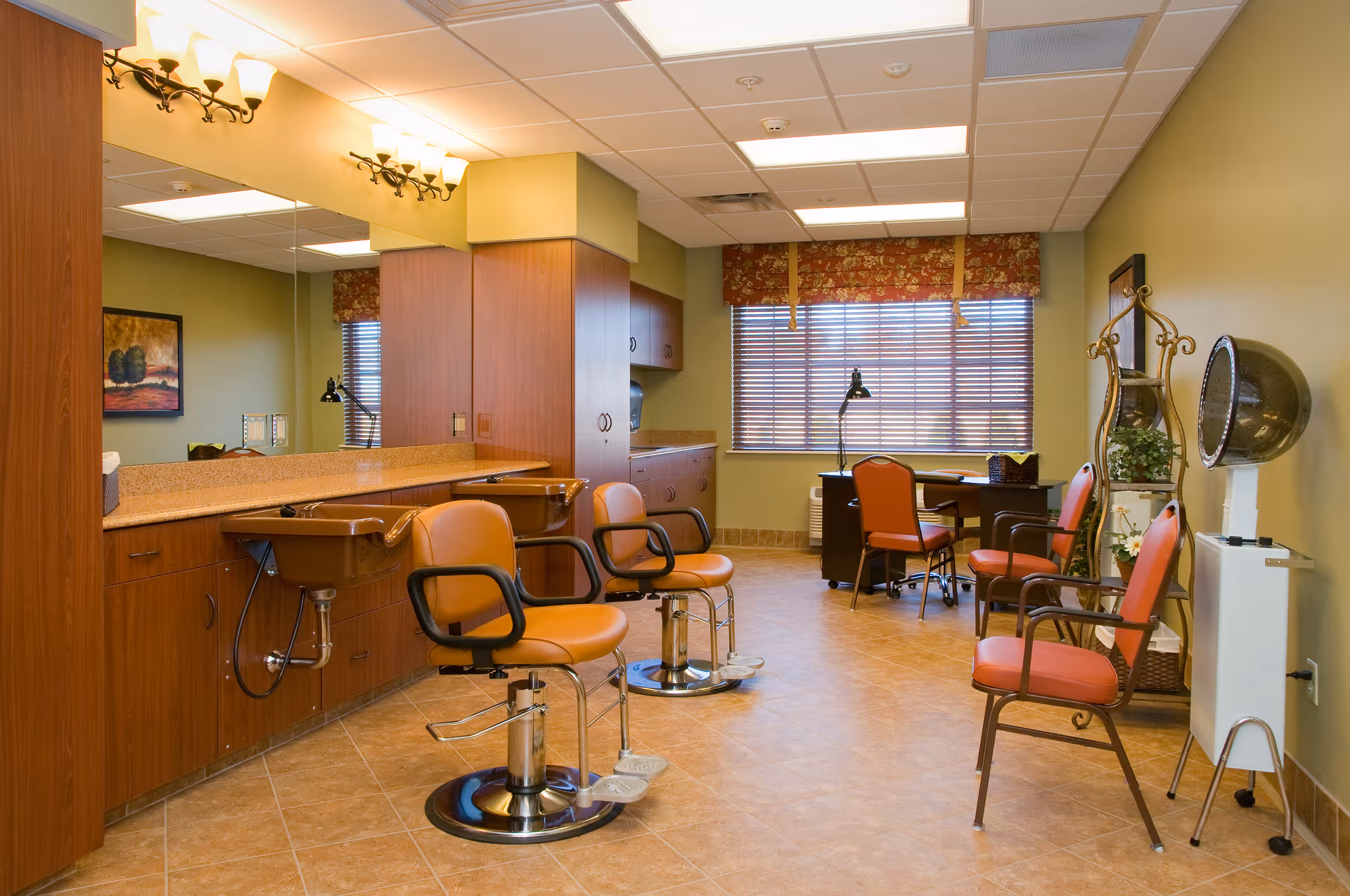 Interior view of a salon area in a senior living facility with two brown salon chairs in front of a long counter with sinks and mirrors. There are additional orange chairs, a desk with a lamp, and a hair dryer station near a window with blinds and floral valance. The room has tiled floors and warm yellow walls.