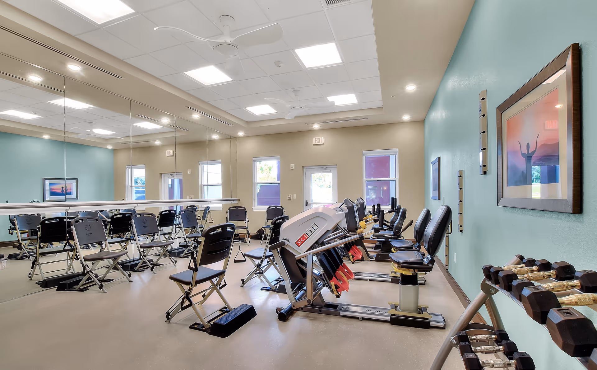 A bright exercise room in a senior living facility with multiple stationary exercise bikes, folding chairs arranged in rows, a large wall mirror, and a rack of dumbbells. The walls are painted light blue and beige, and there are framed pictures on the walls. The room has ceiling lights and windows letting in natural light.