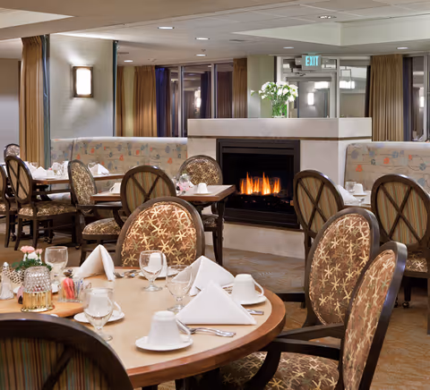 A cozy dining room with round tables set for meals, patterned upholstered chairs, and a central lit fireplace.