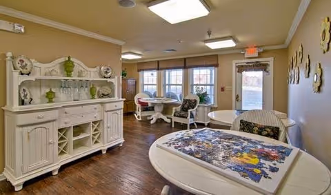 A cozy interior room with wooden flooring featuring white furniture including a sideboard with decorative plates and green vases, round tables with chairs, and a partially completed jigsaw puzzle on one table. The room has large windows with blinds and a door leading outside. The walls are decorated with sunflower wall hangings.