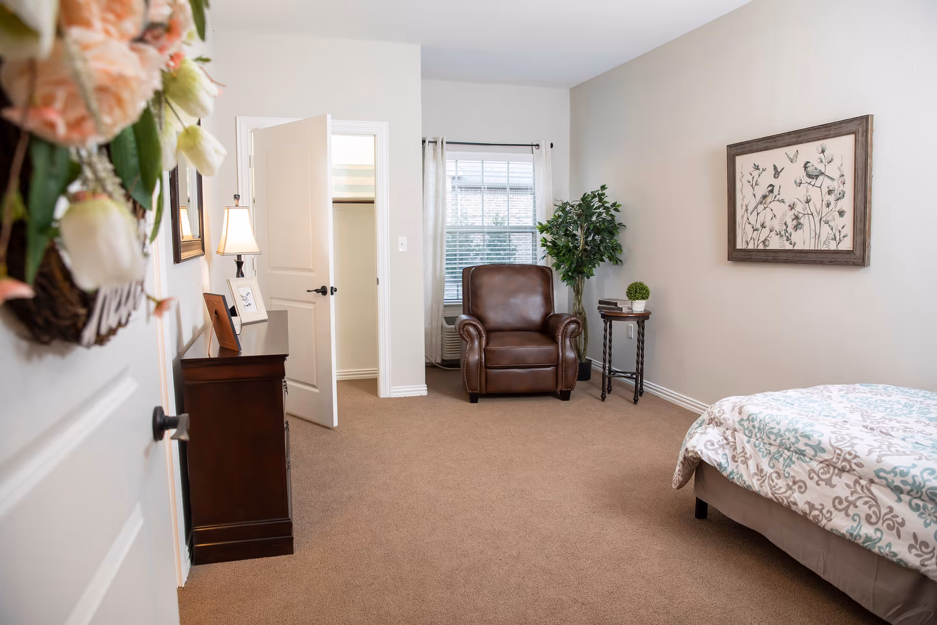 A cozy bedroom with beige carpet and light-colored walls. The room features a bed with a patterned comforter on the right, a brown leather armchair near a window with white curtains, a small side table with decorative items, and a dark wooden dresser with a lamp and framed pictures. A door is open, revealing a closet. A framed artwork of birds and flowers hangs on the wall above the armchair.