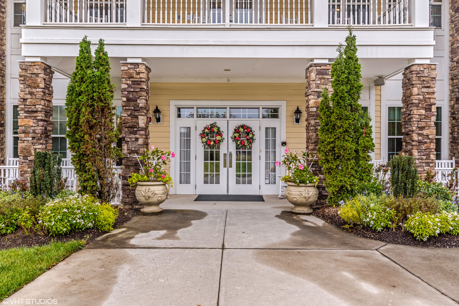 Front entrance of a residential building with double glass doors decorated with wreaths, flanked by stone columns, potted plants, and landscaping.