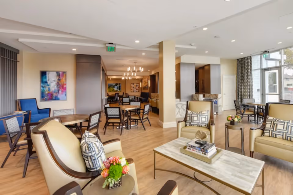 A bright and spacious senior living facility common area with multiple seating arrangements including armchairs with patterned cushions, round wooden tables with chairs, and a coffee table with books and a decorative item. The room features wood flooring, modern lighting fixtures, large windows with patterned curtains, and colorful artwork on the walls.