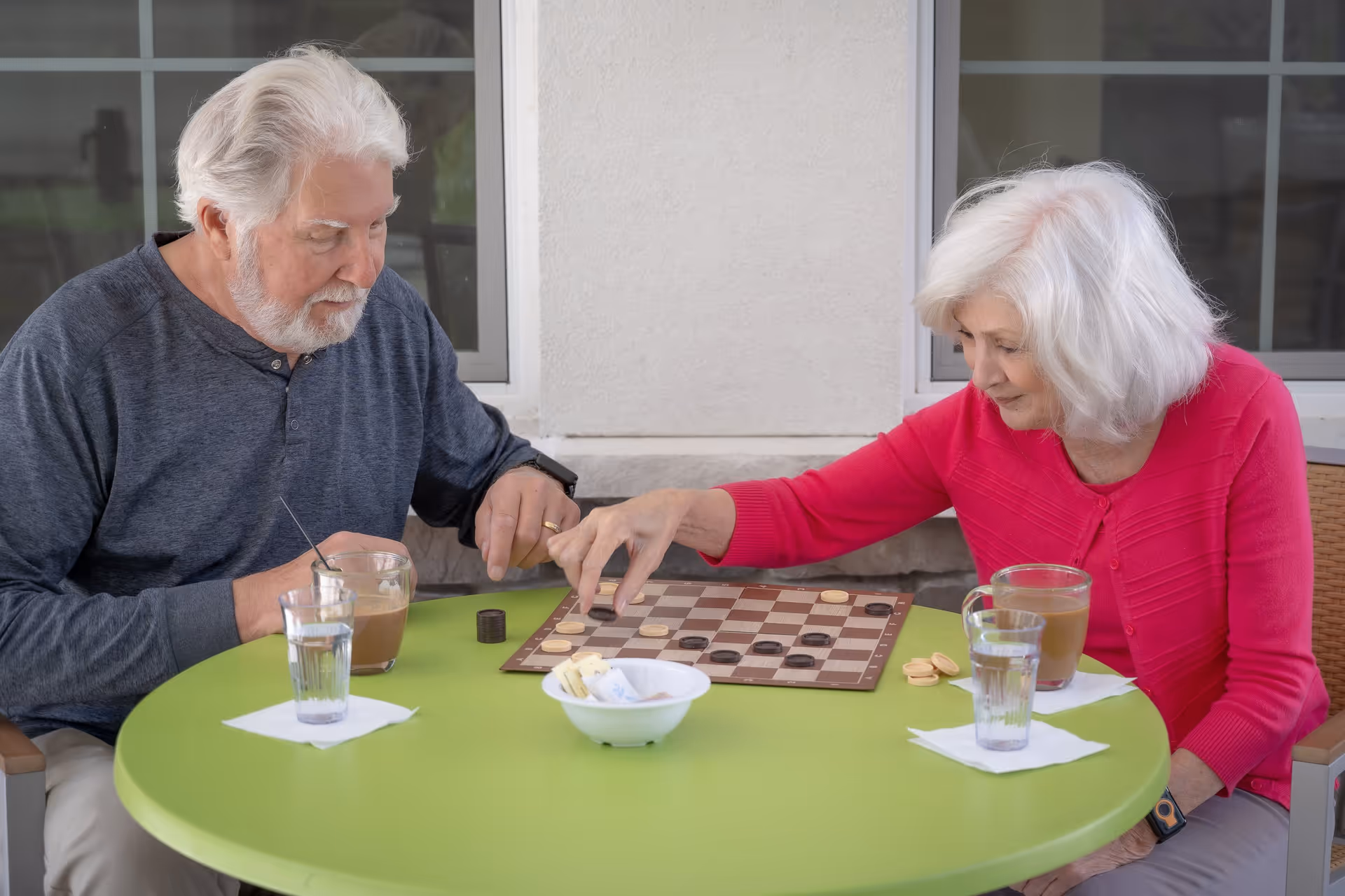 An elderly man and woman sitting at a green round table playing a game of checkers outdoors. Both have white hair; the man is wearing a dark gray long-sleeve shirt and the woman is wearing a bright pink sweater. There are two glasses of water and two cups of coffee on the table, along with a small bowl containing sugar packets.