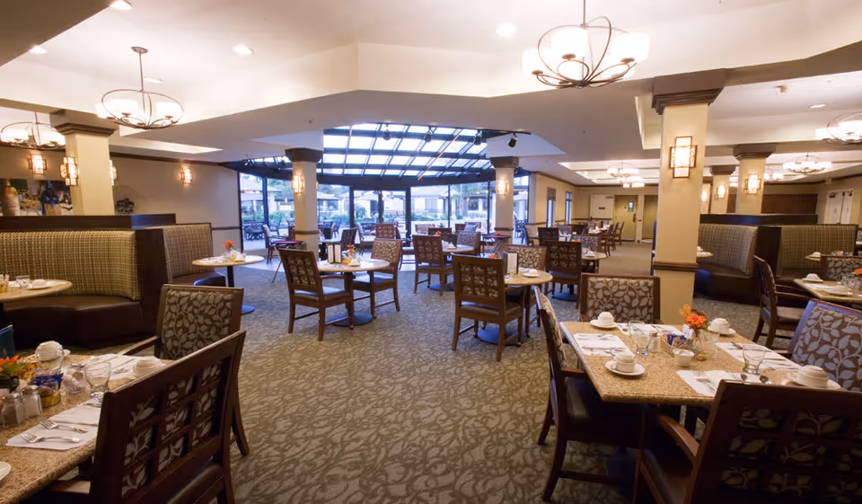 A spacious and well-lit dining room with multiple tables and chairs arranged neatly. The room features patterned carpet flooring, cushioned booth seating along the walls, and decorative light fixtures hanging from the ceiling. Large windows and a glass ceiling section allow natural light to brighten the area, with a view of an outdoor patio space.
