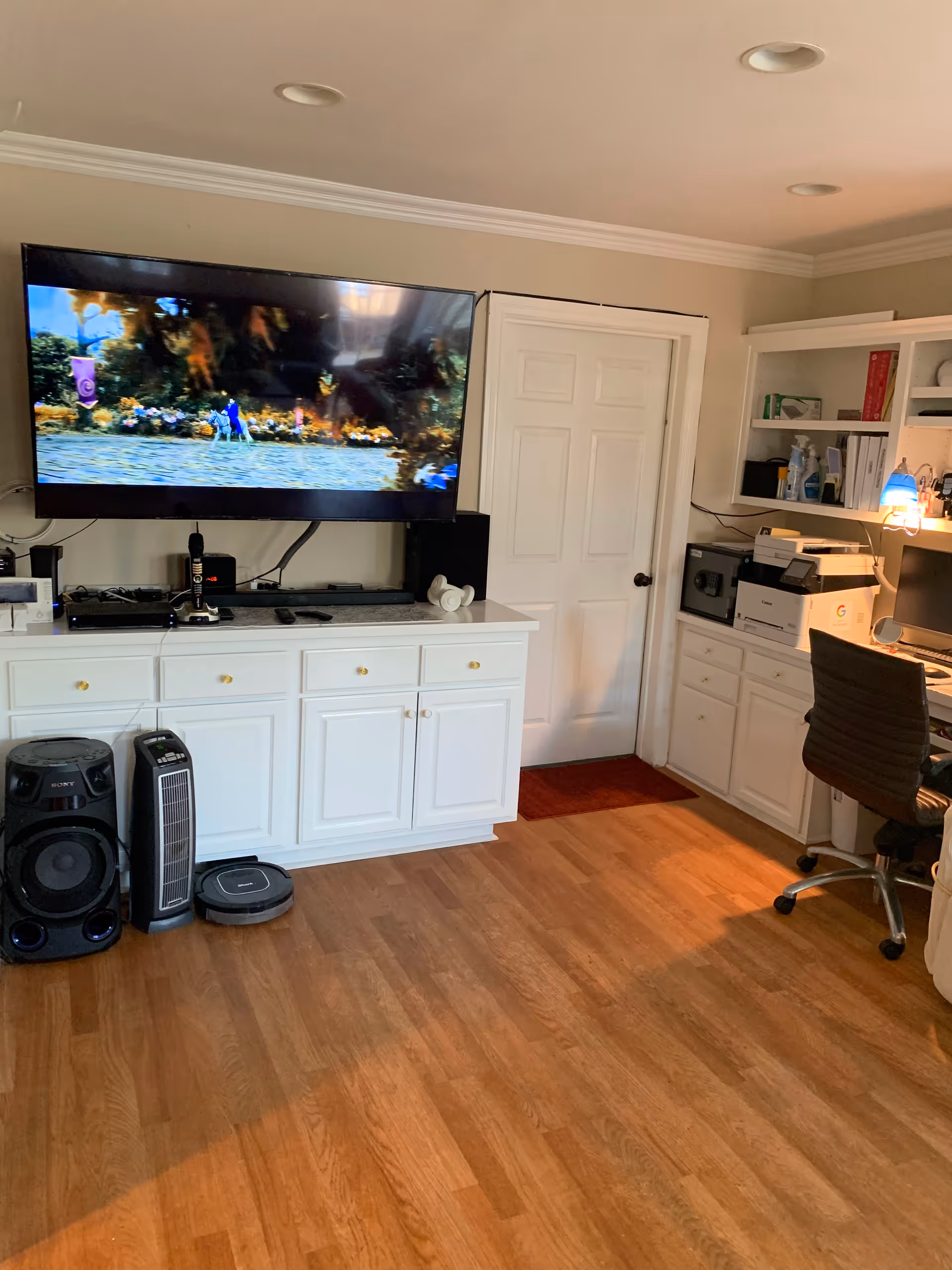 Interior living area with a wall-mounted TV above white cabinets, a desk and office chair to the right, and hardwood floors.