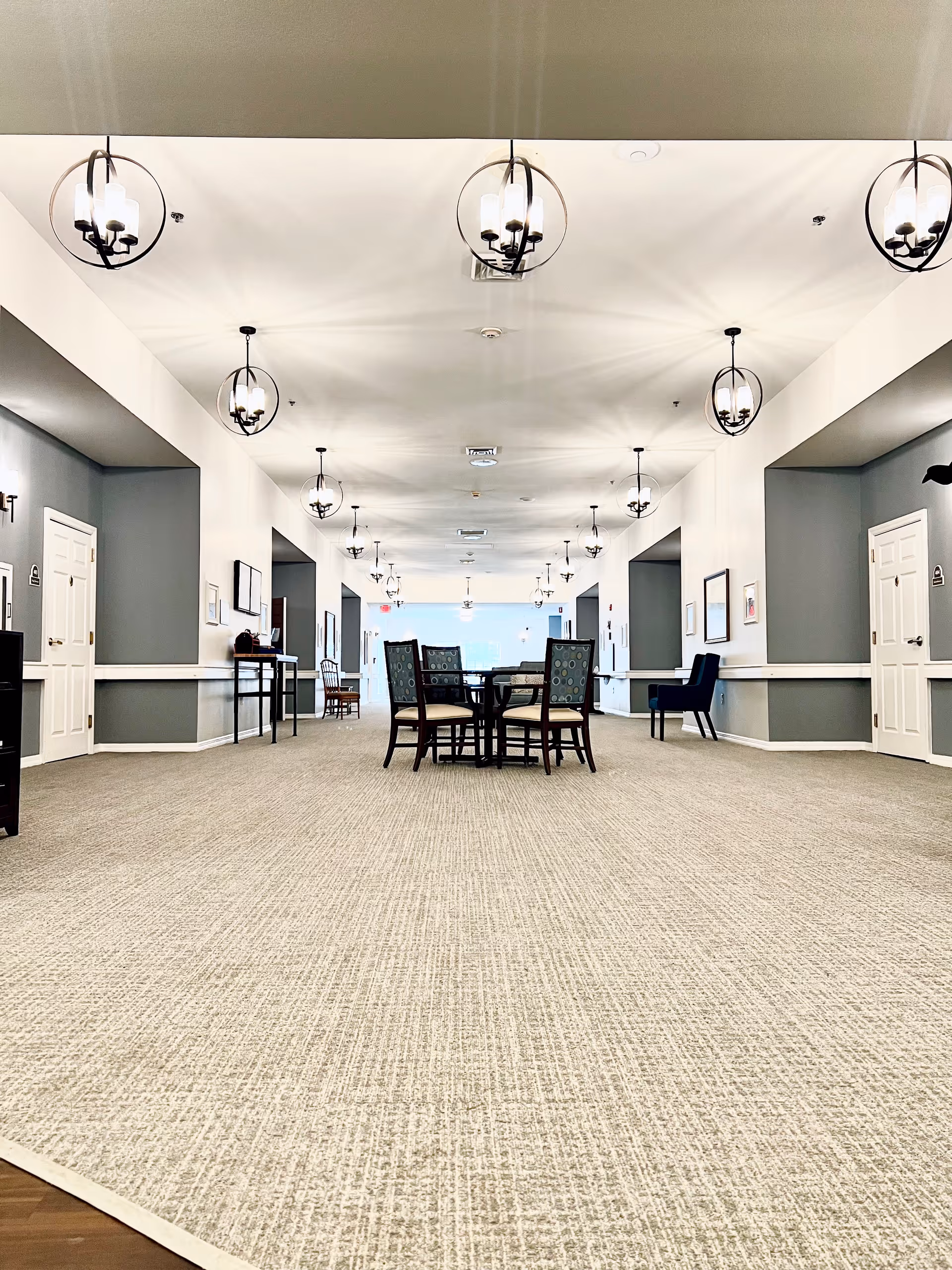 A spacious, well-lit hallway in a senior living facility with beige carpet flooring and white walls accented with gray panels. Several round tables with chairs are arranged along the hallway, and modern circular pendant lights hang from the ceiling. Doors and framed pictures line the walls on both sides.