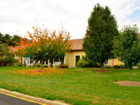 Exterior view of a single-story yellow building with a brown roof, surrounded by green grass and several trees, some with autumn-colored leaves, under a partly cloudy sky.