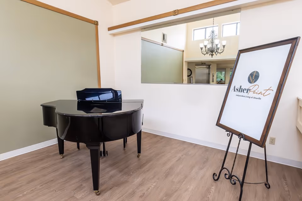 A black grand piano positioned on a light wood floor in a room with white walls and a large window opening to another room. To the right, there is a framed sign on an easel displaying the logo and name 'Asher Point Independent Living of Amarillo'. A chandelier is visible through the window opening.