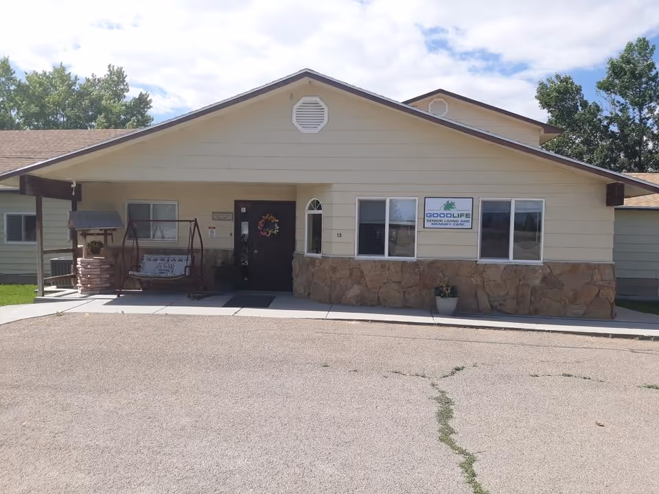 Exterior front view of GoodLife Senior Living & Memory Care building in Elmo, UT. The building has a beige facade with stone accents on the lower half, a dark front door with a decorative wreath, and a porch area with a swing and a small wishing well decoration. There are several windows and a sign displaying the facility's name on the front wall.