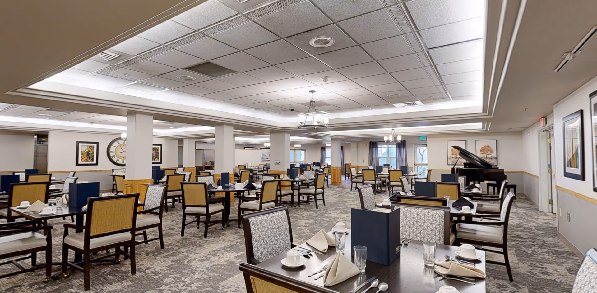 A spacious dining room with multiple tables set for meals, each table featuring folded napkins, cups, glasses, and menus. The room has a patterned carpet, neutral-colored walls with framed artwork, and a grand piano in the corner near windows with curtains. The ceiling has recessed lighting and decorative molding.