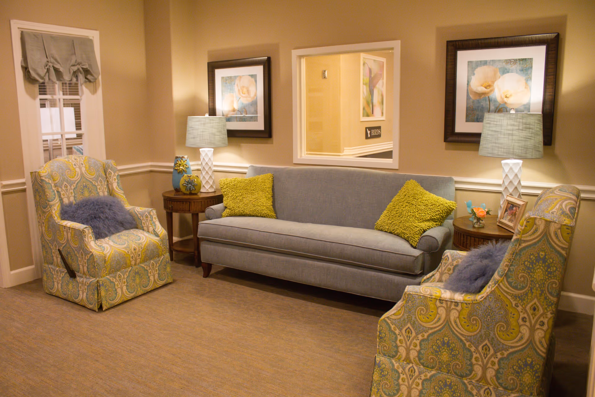 A cozy sitting area with a gray sofa adorned with two green pillows, flanked by two patterned armchairs with blue cushions. There are two wooden side tables, each with a lamp and decorative items. The walls are beige with white trim, and two framed floral artworks hang above the sofa. A window in the wall shows a hallway with a sign reading 'BIRDS'.