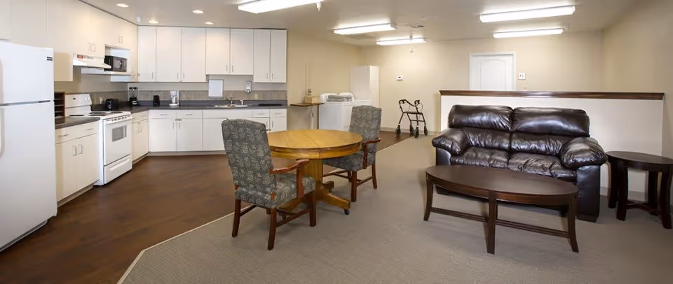 A spacious room combining a kitchen and living area. The kitchen features white cabinets, a white refrigerator, stove, microwave, and various small appliances on the countertop. Adjacent to the kitchen is a round wooden table with two upholstered chairs. The living area includes a dark brown leather sofa, a matching coffee table, and a side table. The floor transitions from wood in the kitchen area to carpet in the living area. A walker is visible near the back wall.