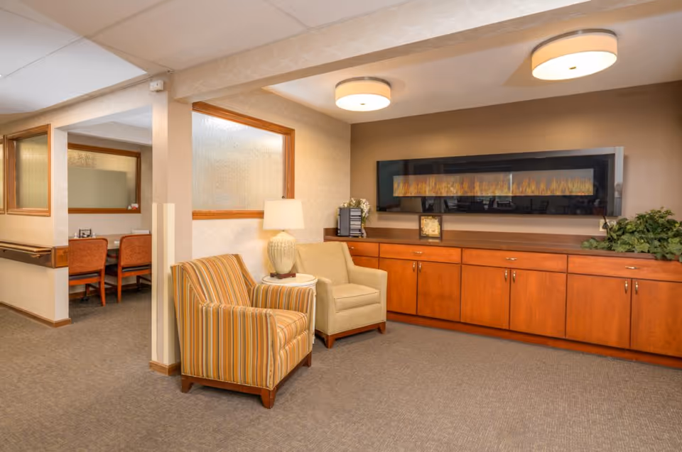 A comfortable common-room seating area with two armchairs, a side table and lamp, wooden cabinets and a long wall-mounted electric fireplace.