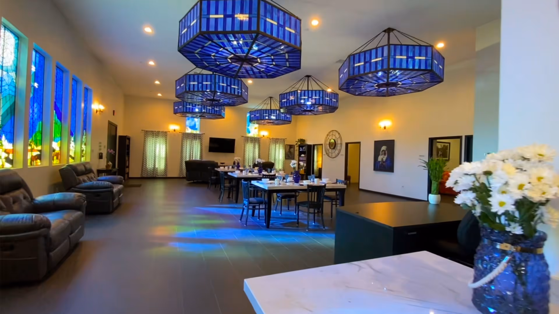 Interior of a senior living facility common area with blue stained glass windows on the left wall, several blue octagonal hanging light fixtures, multiple tables with chairs set for dining, leather recliners along the left wall, and a vase of white flowers on a counter in the foreground.