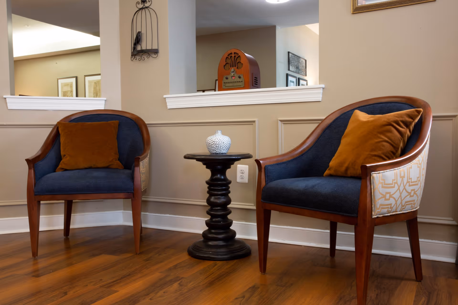 Two wooden armchairs with dark blue cushions and brown throw pillows are positioned on either side of a small black round table with a decorative white vase on top. The setting is indoors with beige walls, wooden flooring, and a partial view of a vintage radio and framed artwork in the background.