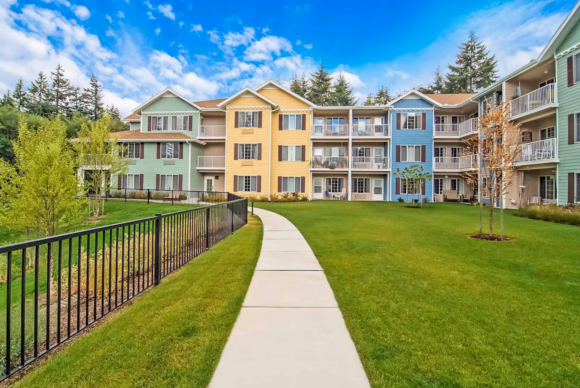 A paved walkway through a green courtyard leads to a colorful three-story senior living building with balconies and landscaping.