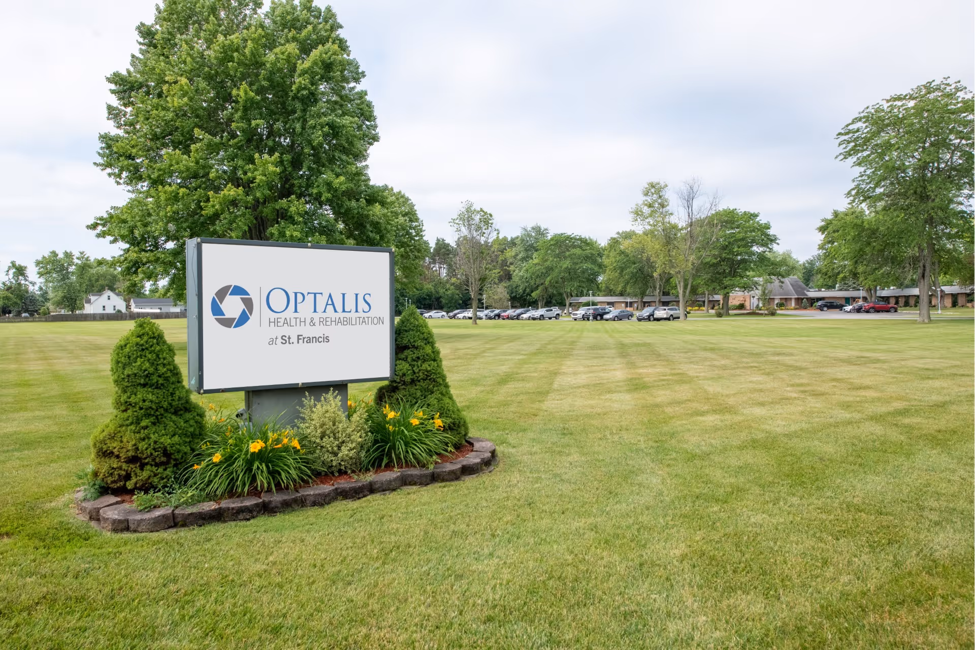 Sign for Optalis Health & Rehabilitation at St. Francis on a manicured lawn with trees and the facility building and parked cars in the background.