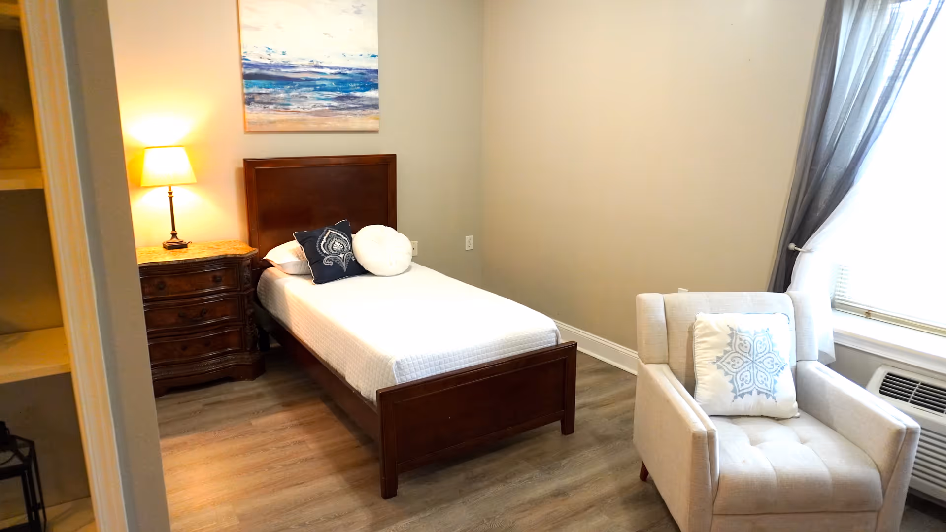 A small bedroom with a single wooden bed with white bedding and two decorative pillows. Next to the bed is a wooden nightstand with a lit lamp. A painting of an ocean scene hangs above the bed. There is a beige armchair with a decorative pillow near a window with sheer curtains and an air conditioning unit below the window. The floor is light wood.