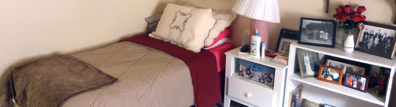 A cozy bedroom featuring a single bed with beige and red bedding, a white nightstand with a lamp, lotion, and books, and a white bookshelf displaying framed photographs and a vase of red flowers.