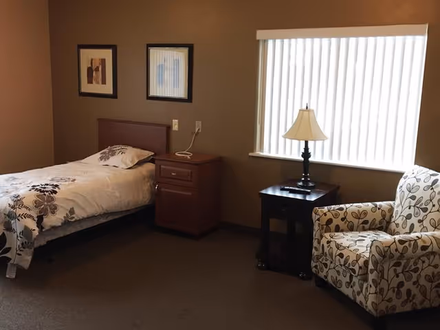 A simple bedroom with a single bed covered in a white and brown floral patterned comforter, a wooden nightstand with a telephone, a window with vertical blinds, a table lamp on a small dark wooden side table, and a cushioned armchair with a leaf pattern.